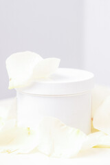 Fototapeta na wymiar White jar with cream among delicate white rose flowers petals on a light background. Natural organic cosmetics concept, close up, mock-up