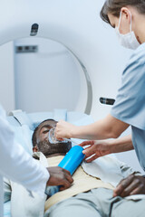 Nurse putting oxygen mask on face of patient lying with closed eyes on MRI table in procedure room