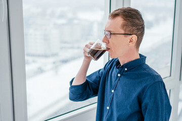 Portrait of focused middle-aged thin man with short ginger hair, glasses in blue long-sleeve shirt looking out through window thoughtfully drinking black coffee from glass cup in morning on balcony