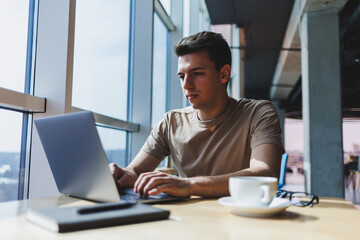 A young attractive male freelancer sits in the interior of a coffee shop and looks out the window. Handsome blogger writing ideas in laptop while sitting at wooden table in cafe with laptop