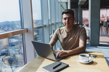 A young attractive male freelancer sits in the interior of a coffee shop and looks out the window. Handsome blogger writing ideas in laptop while sitting at wooden table in cafe with laptop