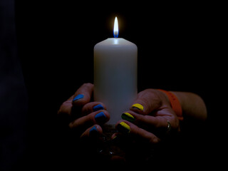 Płomień białej świecy w ciemności trzymanej przez kobietą. Paznokcie kobiety pomalowane są na niebieski i żółty kolor. - obrazy, fototapety, plakaty