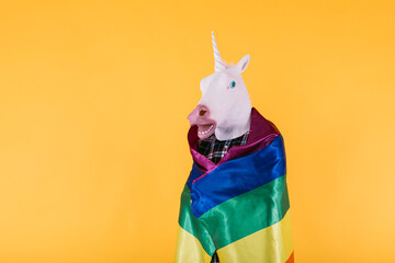 Person dressed in unicorn mask with plaid shirt holding rainbow lgtbq flag, on yellow background. Concept of gay pride, transsexuality, and lgtbq rights.