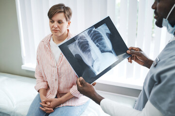 Black doctor analyzing lungs x-ray image of covid patient while talking to female patient about...