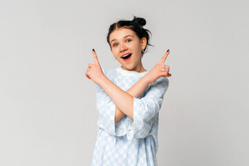 Portrait of indecisive dark-haired teen girl points sideways and smiles, concept of choice, variants, decisions, standing in trendy t shirt over light grey background