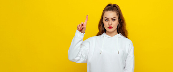 Serious beautiful brunette girl in white casual style sweatshirt, frowning, pointing fingers up, standing over yellow background with copy free space for text