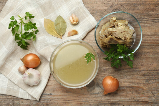 Glass bowl with delicious bone broth and ingredients on wooden table, flat lay