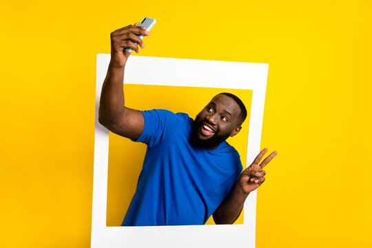 Portrait of handsome trendy cheerful guy coming out of photo frame taking selfie showing v-sign isolated over shine yellow color background