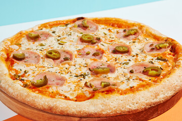 Italian pizza with ham and jalapeno on coloured background. Spicy pizza with jalapeno in minimal style on orange and blue colours. American pizza delivery concept with color backdrop