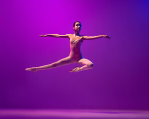 Fototapeta na wymiar Portrait of young little ballet dancer, teen jumping isolated on purple background in neon light. Art, grace, beauty, ballet school concept