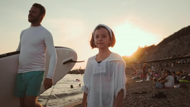 Surfing. Father with sup board and daughter are walking along the beach. Dolly shot, Slow motion. Sunset on the background.