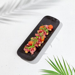 Roast beef with cherry tomatoes, ruccola and spicy sauce on black plate. Sliced roast beef on white concrete background. Italian antipasti in summer style with hard shadow and palm leaves.