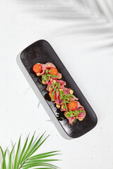 Roast beef with cherry tomatoes, ruccola and spicy sauce on black plate. Sliced roast beef on white concrete background. Italian antipasti in summer style with hard shadow and palm leaves.