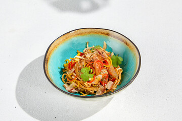 Stir fry noodles with chicken and bacon in asian style. Fried udon with meat and vegetables in ceramic bowl on white background and sunlights shadows. Asian food in modern style.