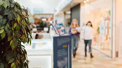 Abstract commercial background. Defocused store, blurred display windows with advertising and buyers people walking along corridor of supermarket. Selective focus on green plant
