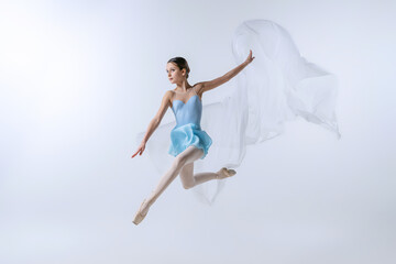 Fototapeta premium One young girl, aspiring ballerina in blue dress and pointes dancing with cloth isolated on gray background. Art, grace, beauty, ballet school concept