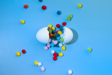 Fototapeta na wymiar Cracked white egg with multi-colored round pom-poms falling out of it on a blue background, top view