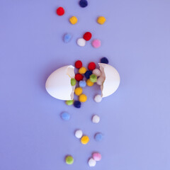 A cracked white egg with multi-colored round pom-poms falling out of it on a purple background, top view