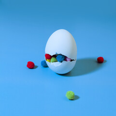 A white egg with a big crack and colorful pom-poms inside on a blue background, an egg with a surprise