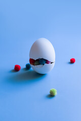 A white egg with a big crack and colorful pom-poms inside on a blue background, an egg with a surprise