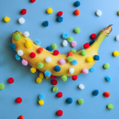 Funny banana with pom-poms, a banana covered with round colored pom-poms on a blue background and surrounded by scattered pom-poms, strange fruit concept