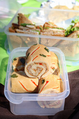 A bread roll with tuna filling mixed with mayonnaise and onion, coriander and red bell pepper in a lunch box on a mat with club sandwiches in background with bright sunshine 