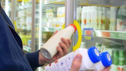 An unidentified man in a store selects products at the dairy counter, reads the composition and checks the expiration date, and then puts the purchases in a basket for payment at the checkout.