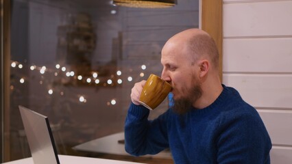 A bald man with a beard sits at a table working remotely from home after his main job in order to feed his family. A middle-aged man drinks coffee at home while working at a computer.