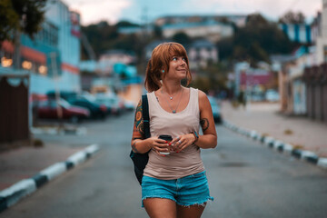 Young smiling woman with tattoo walking with a cup of coffee in her hands. Evening stroll
