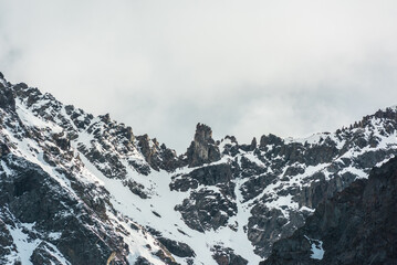 Fototapeta na wymiar Dramatic landscape with high snowy mountain with peaked top in cloudy sky. White snow on black sharp rocks. Awesome snow mountain range in changeable weather. Sharp pointed peak with snow in cloud sky