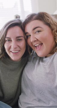 Vertical shot of a Caucasian lesbian couple having a video call, smiling at the camera and holding k