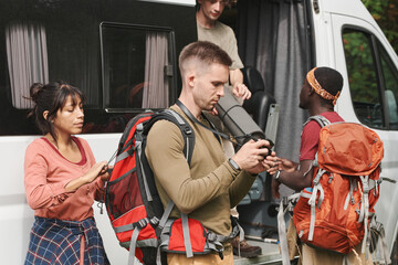 Serious young man looking through photos on camera display while his friends unloading hiking stuff from van