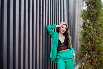 Smiling woman in green suit and black delicate top posing next to black rack wall outdoor.