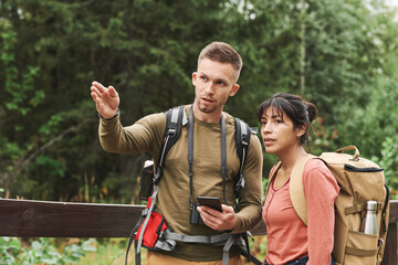 Young Caucasian hiker with camera hanging on neck gesturing hand and using phone while explaining route to woman