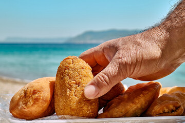 Typical Sicilian street food on seaside background. Hand of man taking arancina (deep fried rice...