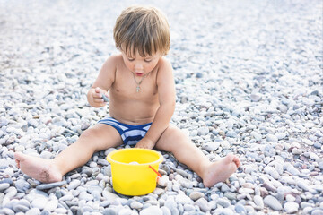 Cute child playing with yellow bucket and toy car on pebble beach. Happy toddler on sea coast. Summer family holiday vacation. Two years old little boy having fun. Summertime. Travel concept