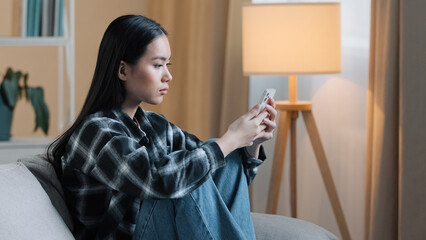 Fototapeta na wymiar Side view of sad bored Asian woman girl sitting on sofa at home with telephone serious unhappy lady scrolling browsing in smartphone and using mobile phone app watching social media news feed typing