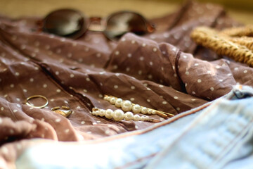 Brown polka dot blouse, vintage mom jeans, straw bag, round sunglasses, gold rings and pearl hair clips. Fashionable outfit on wooden background. Selective focus.