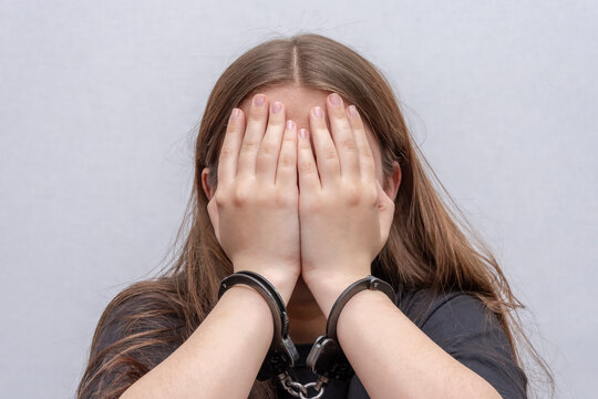 A young girl handcuffed hides her face on a gray background, close-up. Juvenile delinquent in a black T-shirt, criminal liability of minors.