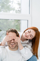 Smiling woman covering eyes of boyfriend near window at home.