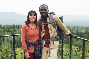 Fototapeta premium Happy multi-ethnic couple standing at railings against forest landscape and looking at camera at hike