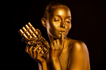 Photo of gentle lady body painted gold shimmer eyes closed touch her lip hold chocolate bar...