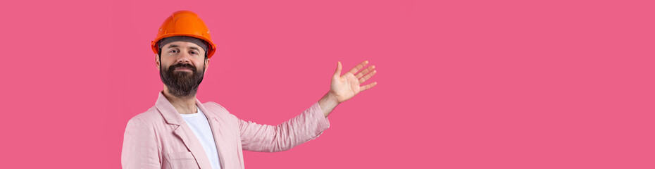 Portrait of young construction engineer wear orange hard hat, in a pink jacket standing on red studio background. The man points with his hand.