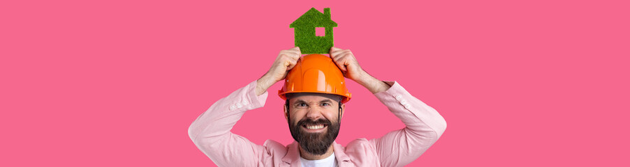 Portrait of young construction engineer wear orange hard hat, in a pink jacket standing on red studio background. A man holds a green eco house.