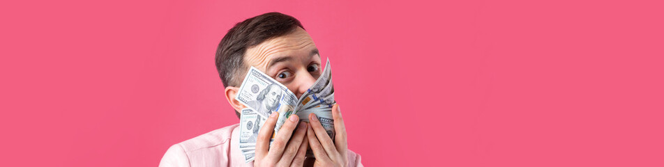 Portrait of a contented young businessman with a beard dressed in a pink jacket showing us dollar banknotes against a red studio background. Taste, smell of money.