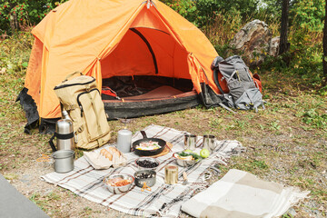 Various dishes cooked on burner placed on cloth against orange tent and backpacks in forest