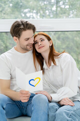 Positive man holding card with blue and yellow heart sign and looking at girlfriend at home.