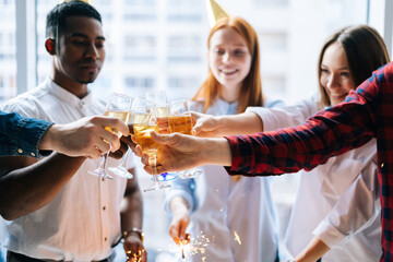 Close-up cropped shot cheerful business team of young multi-ethnic businesspeople holding Bengal lights and clinking glasses of champagne at corporate party, on background of window and cityscape.