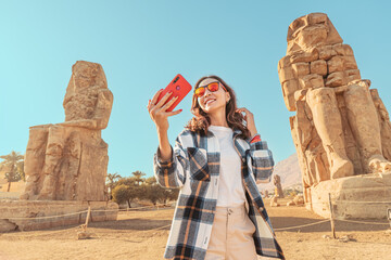 Travel blogger girl takes selfie pictures on a smartphone at the famous two Colossi of Memnon -...