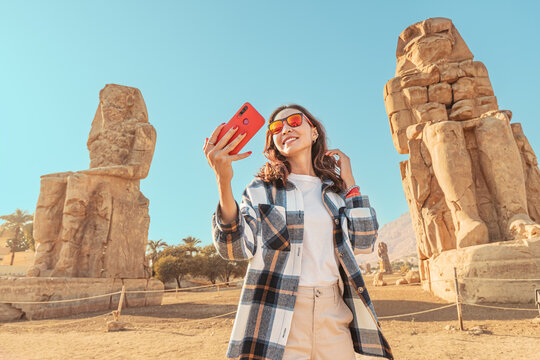 Travel blogger girl takes selfie pictures on a smartphone at the famous two Colossi of Memnon - massive ruined statues of the Pharaoh Amenhotep III. World tourism attractions